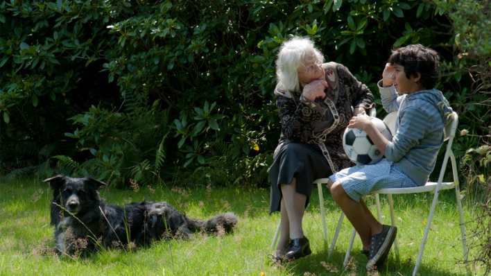 Ein Junge mit Ball und eine ältere Frau sitzen auf Stühlen nebeneinander und unterhalten sich. Daneben liegt ein schwarzer Hund (Quelle: Joachim M. Huber)