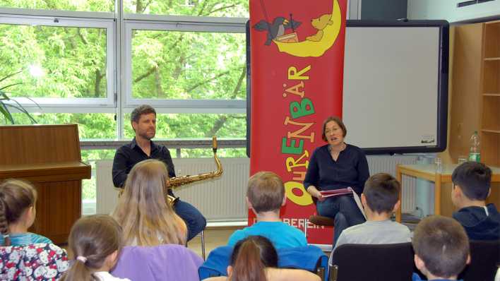 Autorin Ariane Grundies liest vor Schülern in einem Bibliotheksraum, links daneben sitzt Musiker Tilman Ehrhorn mit Saxophon (Quelle: rbb/OHRENBÄR/Birgit Patzelt)