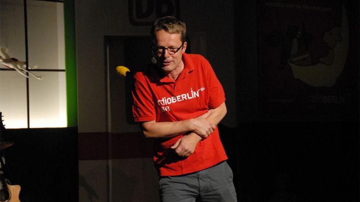 radioBERLIN-Moderator Alexander Schurig auf der Bühne, hält sich den linken Arm (Quelle: rbb/OHRENBÄR/Birgit Patzelt)