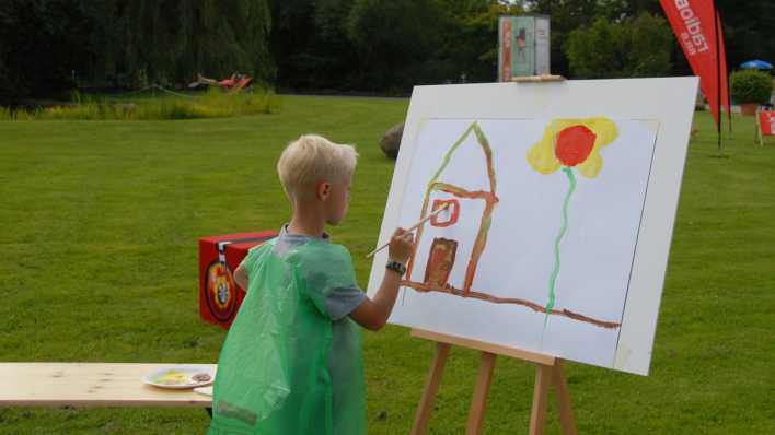 Ein Junge malt ein braunes Haus, daneben hat er schon eine große Blume gemalt, an der Staffelei (Quelle: rbb/OHRENBÄR/Birgit Patzelt)