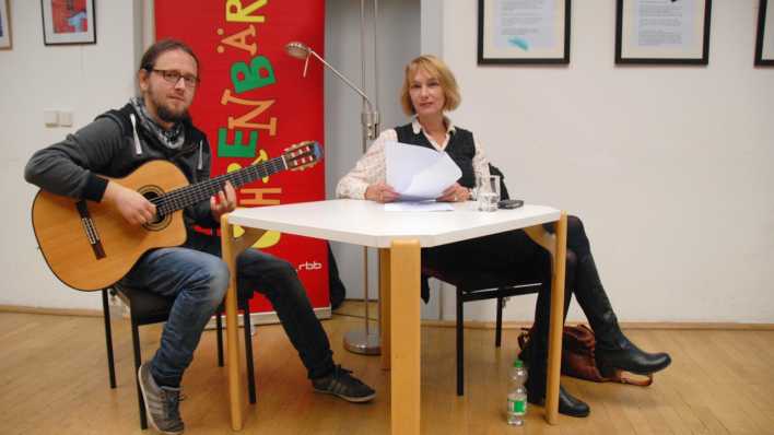 Schriftstellerin Nina Petrick und der Gitarrist Jacob Jurgeit am Lesetisch, im Hintergrund ein OHRENBÄR-Banner (Qoelle: rbb/OHRENBÄR/Birgit Patzelt)