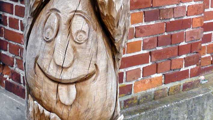Geschnitzte Holzfigur an einer Häuserwand, sie lacht und streckt die Zunge raus (Quelle: rbb/OHRENBÄR/Sonja Kessen)