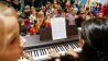 Eine Frau spielt auf einem E-Piano, daneben ein Mädchen, davor viele Kinder, die mitsingen (Quelle: Klax Schule/Frederike Coring)