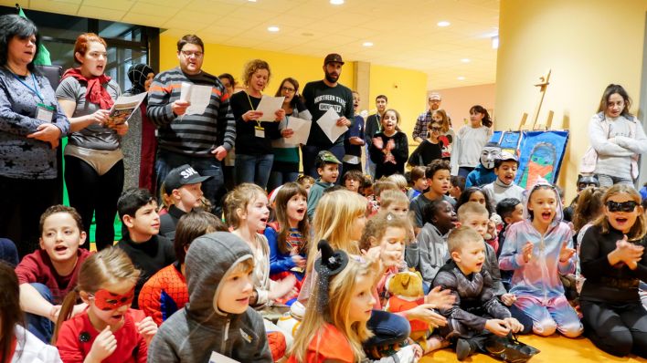 Viele Kinder sitzen auf dem Boden, dahinter Erwachsene, alle haben einen Textzettel und singen (Quelle: Klax Schule/Frederike Coring)