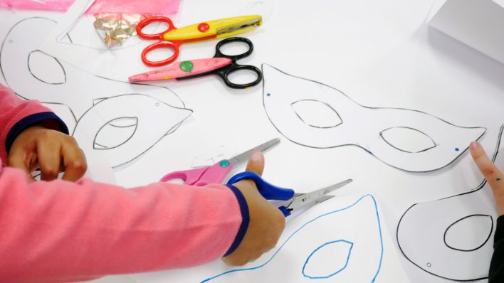 Augenmasken auf einem großen weißen Blatt gemalt, Kinderhände schneiden mit der Schere die Masken aus (Quelle: Klax Schule/Frederike Coring)