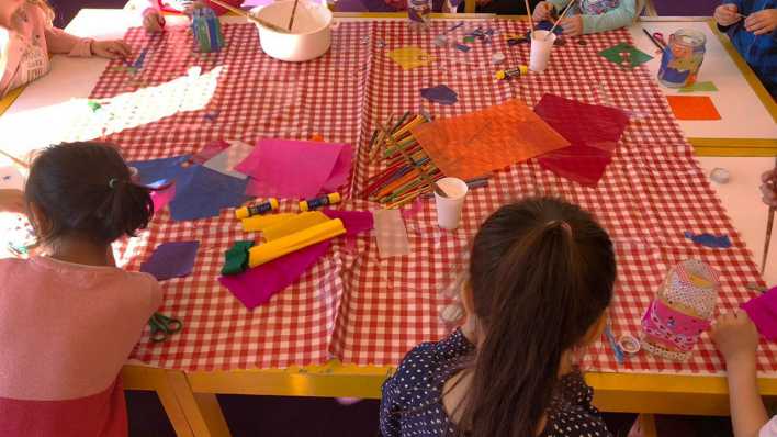 Ein großer Tisch mit rot-weiß karierter Decke, drumherum viele Kinder, darauf Bastelmaterialien (Quelle: rbb/OHRENBAER/Kathi Heidel)