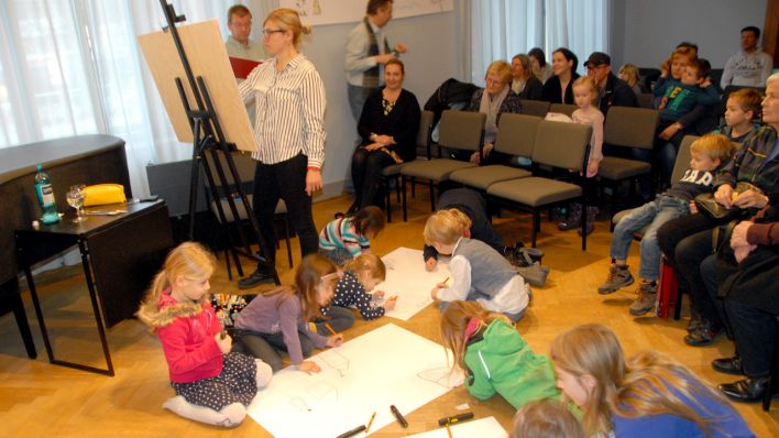 Sandra Bach mal live zur Geschichte, im Hintergrund OHRENBÄR-Autor Hubert Schirneck liest, davor: Kinder malen auf Blätter, die auf dem Boden liegen (Quelle: rbb/OHRENBÄR/Birgit Patzelt)