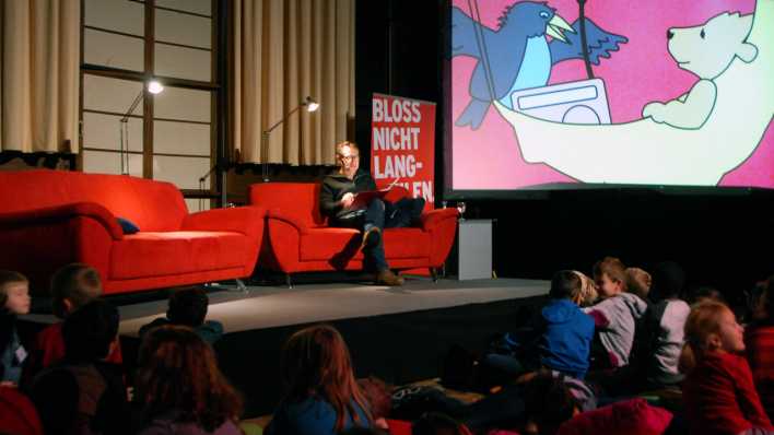 Schauspieler Boris Aljinovic auf dem roten Sofa, im Hintergrund das OHRENBÄR-Logo (Quelle: rbb/OHRENBÄR/Birgit Patzelt)