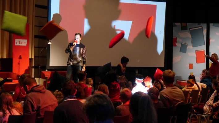 rbb-Moderator Marc Langebeck auf der Bühne, kissen fliegen in der Luft, viele Kinder und wenige Erwachsen im Publikum (Quelle: rbb/OHRENBÄR/Sonja Kessen)