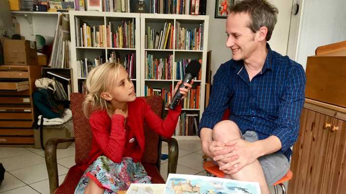 Kinder-Reporterin Lientje interviewt Illustrator Jens Rassmus, im Hintergrund ein großes Bücherregal (Quelle: rbb/OHRENBÄR/Janine Lüttemann)