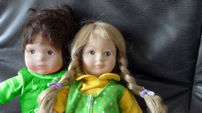 Zwei Puppen, Junge und Mädchen, mit grün-gelber Puppenkleidung auf einer dunklen Ledercouch (Quelle: Karin Gähler)