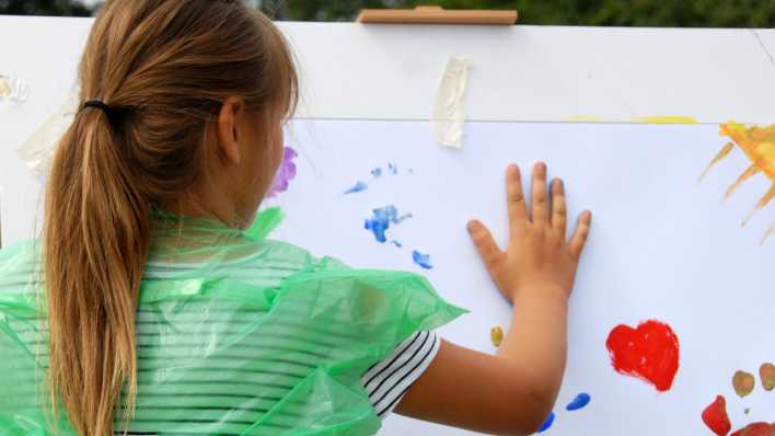 Ein Mädchen mit Zopf an einer Staffelei, sie stempelt mit der rechten Hand Farbe auf ein bemaltes Blatt (Quelle: rbb/OHRENBÄR/Birgit Patzelt)