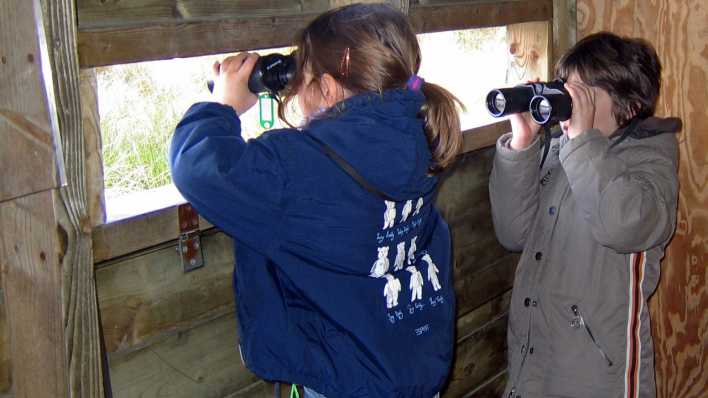 Ein Mädchen und ein Junge beobachten mit Ferngläsern die Umgebung, aus einem Holzschuppen (Quelle: imago images/Blickwinkel)