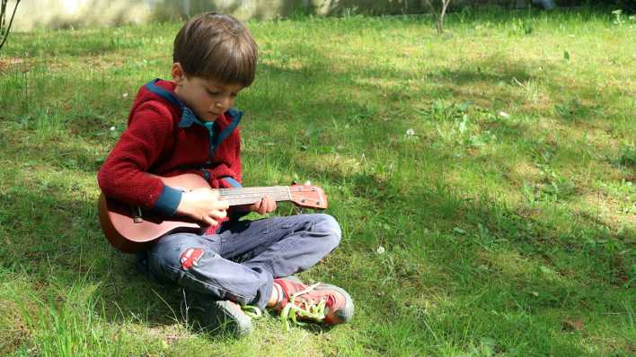 Ein Junge mit Jeans und roter Kapuzenjacke: sitzt auf der Wiese und spielt Ukulele (Quelle: Martha Zan)