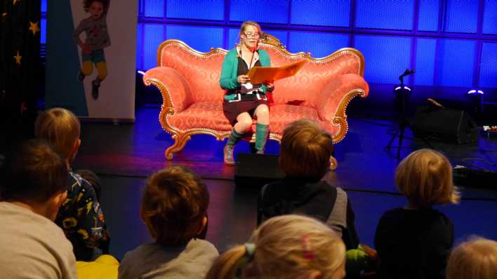 Schauspielerin Astrid Kohrs auf der Bühne, sie sitzt auf einer eleganten Couch und liest OHRENBÄR-Geschichten (Quelle: rbb/OHRENBÄR/Sonja Kessen)