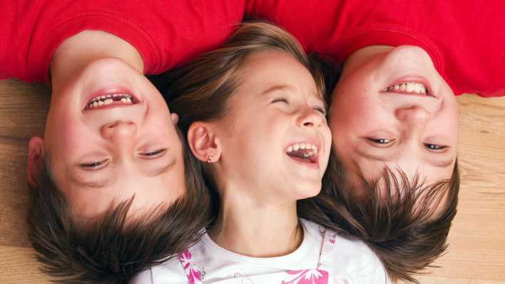 Drei fröhliche Kinder, ein Mädchen und zwei Jungen, liegen auf dem Boden, Köpfe aneinander (Quelle: imago images/imagebroker)