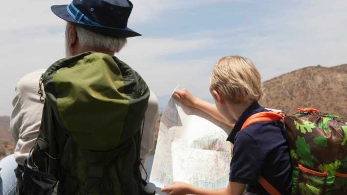Opa und Enkel sitzen vor bergiger Landschaft, mit Rucksäcken, Opa trägt einen Hut, der Enkel schaut auf eine Karte (Quelle: imago images/ingimage)