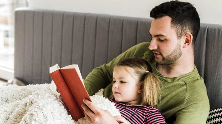 Vater liest seiner Tochter vor, auf einer Couch, mit weißer Kuscheldecke (Quelle: imago images/Westend 61)