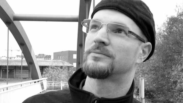 Autorenfoto von Andreas Hartmann, schwarz-weiß, mit Brille und Mütze, auf einer Brücke (Quelle: privat)