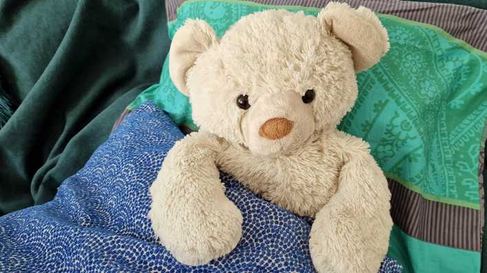 Ein Teddybär, eingekuschelt in eine blau-weiße Decke (Quelle: rbb/OHRENBÄR/Sonja Kessen)
