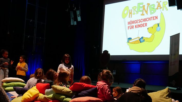 OHRENBÄR-Logo auf großer Leinwand, davor kuscheln sich Kinder in die Kissen (Quelle: rbb/OHRENBÄR/Sonja Kessen)
