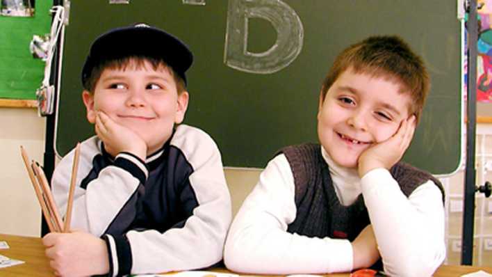 Zwei Jungs, Hand aufgestützt, sitzen vor einer Tafel, auf der Kreidebuchstaben sind, und lächeln (Quelle: imago/Reinhard Kunzendörfer)