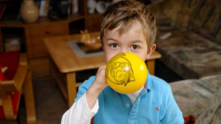 Kleiner Junge mit blauem Shirt bläst einen gelben Luftballon auf (Quelle: imago/Torsten Becker)