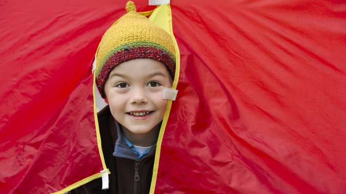 Kleiner Junge mit gelber Mütze schaut durch einen roten Zelteingang (Quelle: imago/Torsten Becker)