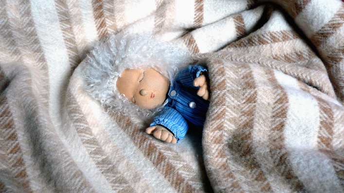 Eine kleine Puppe mit Struwelhaaren und blauer Jacke schläft in einer weiß-braunen Wolldecke (Quelle: rbb/OHRENBÄR/Sonja Kessen)