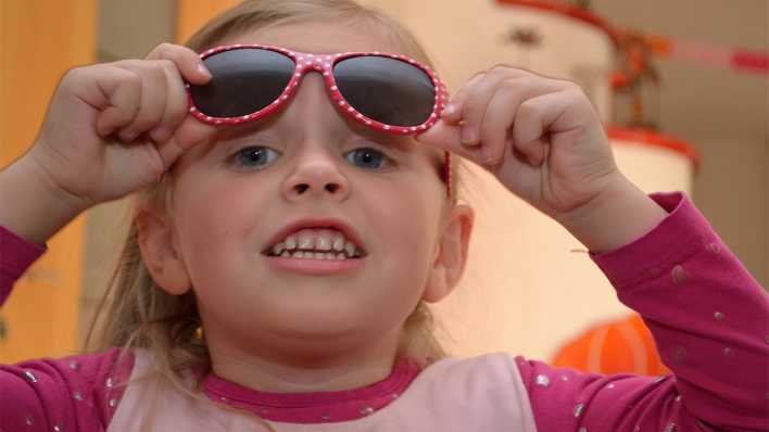 Ein blondes Mädchen mit rosaroter Sonnenbrille, beide Hände an der Brille (Quelle: imago/CHROMORANGE)