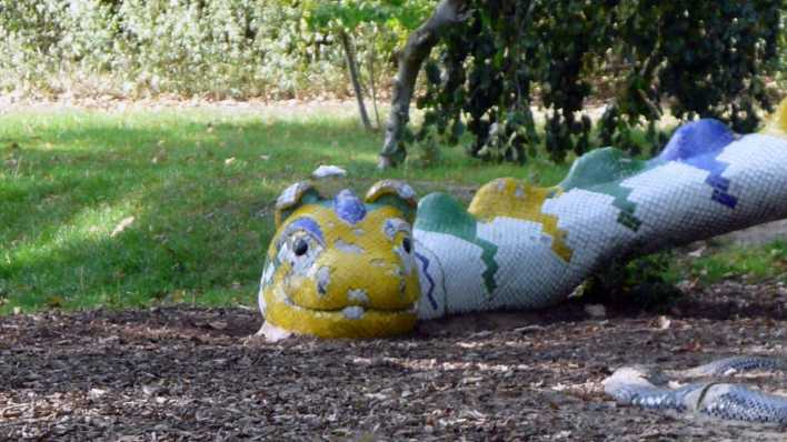 Auf einer Wiese, im Hintergrund Büsche/Bäume, liegt ein bunter Drache aus Holz (Quelle: rbb/OHRENBÄR/Sonja Kessen)