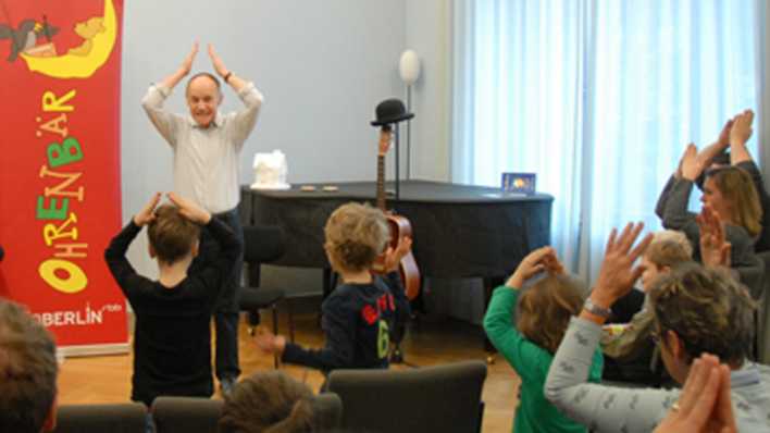 Musiker Robert Metcalf macht mit den Händen über seinem Kopf eine Zipfelmütze, alle im Saal machen mit, Blick zur Bühne. © rbb/OHRENBÄR/Birgit Patzelt