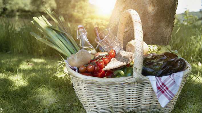 Ein gefüllter Picknickkorb auf einer grünen Wiese, im Hintergrund ein Baum, die Sonne geht unter (Quelle: imago images/Westend61)