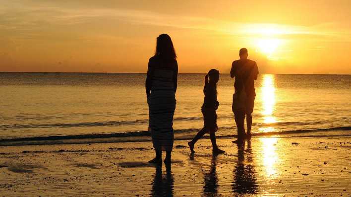 Dämmerung, eine Familie (Frau, Kind, Mann) am Meeresstrand, die Sonne geht unter (Quelle: imago images/EQ Images)