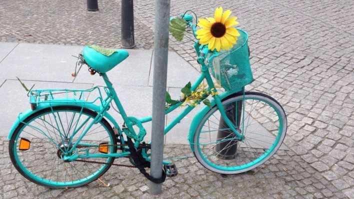 Ein mintfarbenes Fahrrad an einem Straßenpfeiler angeschlossen, im Korb am Lenker ist eine große Sonnenblume (Quelle: rbb/OHRENBÄR/Birgit Patzelt)