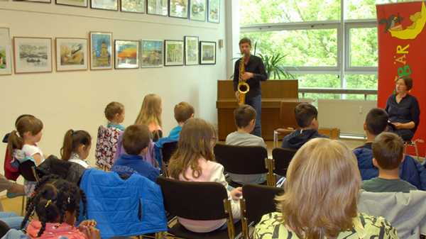 Tilman Ehrhorn spielt vor Kindern in einem Bibliotheksraum Saxophon, rechts sitzt Autorin Ariane Grundies (Quelle: rbb/OHRENBÄR/Birgit Patzelt)