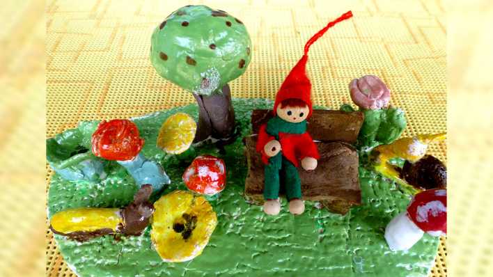 Gebrannte Keramik: ein Wichtel mit roter Zipfelmütze auf einer Bank im Garten, mit Schnecken, Baum, Pilzen und Blumen (Quelle: Karin Gähler)