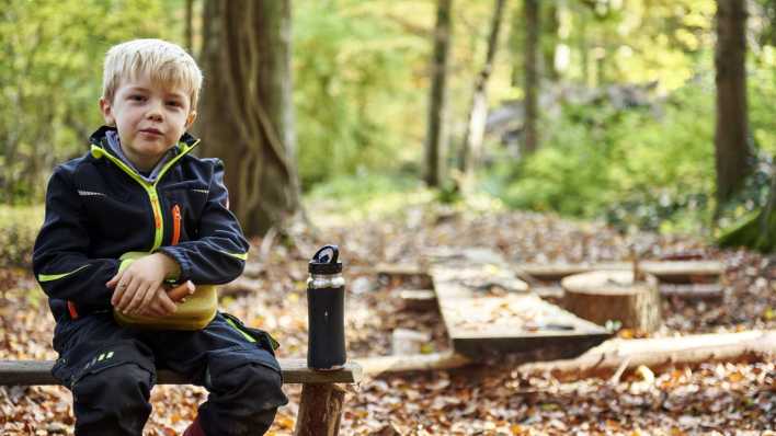 Ein blonder Junge mit Würstchen, Brotbüchse und Trinkflasche auf einer Holzbank, im Hintergrund Mischwald (Quelle: imago images/Westend61)