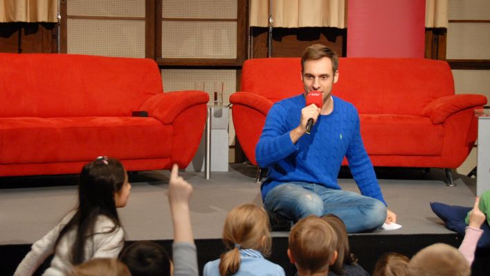 Moderator Marc Langebeck sitzt auf dem Bühnenboden, hält ein Mikro in der Hand, vor ihm Kinder, einige melden sich (Quelle: rbb/OHRENBÄR/Birgit Patzelt)
