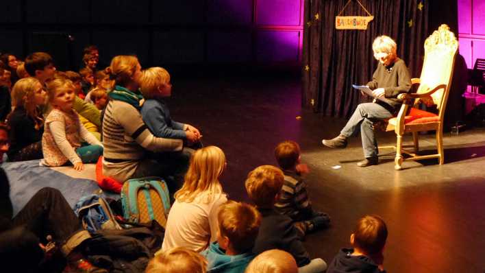 OHRENBÄR-Autorin Gudrun Mebs im Lesesessel auf der Bühne, Kinder und Erwachsene im Publikum (Quelle: rbb/OHRENBÄR/Sonja Kessen)