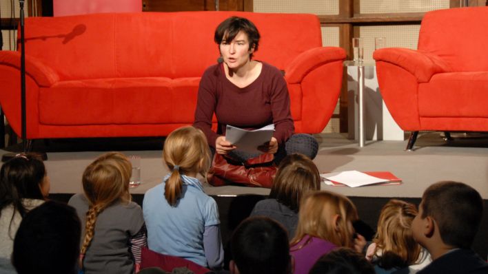 Autorin Christine Anlauff sitzt auf der Bühne, vor ihr liegen aufgeschlagene Bücher, sie spricht zu den Kindern vor der Bühne (Quelle: rbb/OHRENBÄR/Birgit Patzelt)