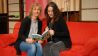 Nina und Conni von Zappelduster sitzen auf der roten Bühnencouch, halten Buch und Mikrofon in den Händen (Quelle: rbb/OHRENBÄR/Birgit Patzelt)