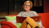 Schauspielerin Karen Matting sitzt auf der roten Lesecouch, liest und blickt ins Publikum (Quelle: rbb/OHRENBÄR)