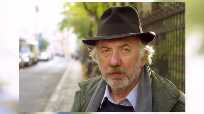 Autor Helmut Eisendle mit Hut und Schal auf einer Straße, im Hintergrund ein Zaun (Quelle: imago images/gezett)