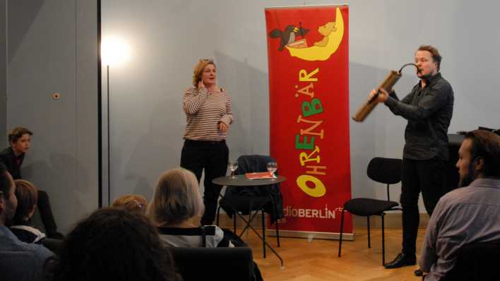 im Vordergrund die Köpfe des Publikums, im Hintergrund Karen Matting steht am Tisch und Friedemann Matzeit spielt Melodica (Quelle: rbb/OHRENBÄR/Birgit Patzelt)