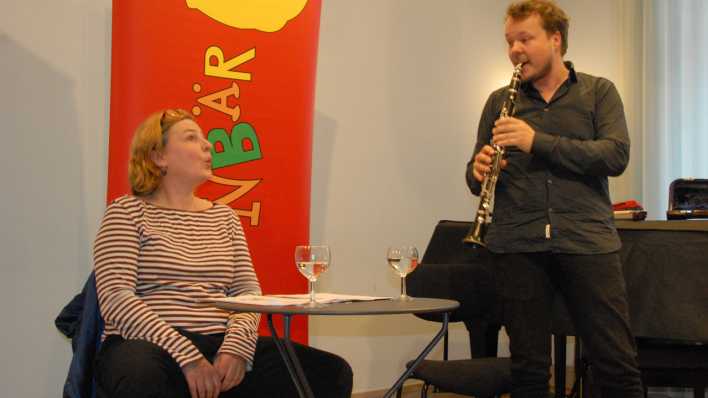 Karen Matting sitzt am Tisch und Friedemann Matzeit spielt Klarinette, im Hintergrund das rote OHRENBÄR-Banner (Quelle: rbb/OHRENBÄR/Birgit Patzelt)