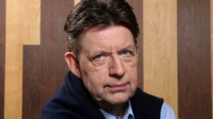 Porträt des Schauspielers Jörg Gudzuhn vor einer braunen Holzwand, blauer Pullover über den Schultern (Quelle: imago images/Sven Simon) Porträt des Schauspielers Jörg Gudzuhn vor einer braunen Holzwand, blauer Pullover über den Schultern (Quelle: imago images/Sven Simon)