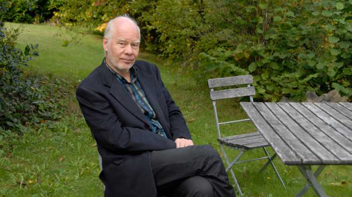 Porträt des Autors Lukas Hartmann, sitzend an einem Tisch im Garten (Quelle: Regine Mosimann / © Diogenes Verlag)