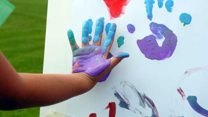 Eine bunt bemalte Kinderhand, vor der Staffellei mit bunten Kinderhandabdrücken (Quelle: rbb/OHRENBÄR/Birgit Patzelt)