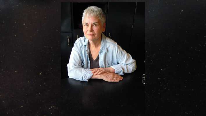 Porträt der Autorin Hanna Johansen, Arme auf einem schwarzen Tisch, im Hintergrund ein schwarzer Schrank (Quelle: privat)